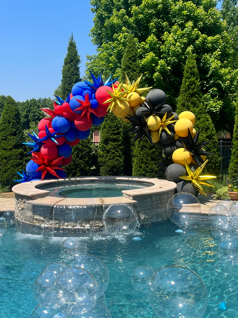 Balloon arch over a outdoor pool, half red and blue, half black and gold, clear balloons float on pool like bubbles