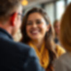 Woman in yellow scarf smiling at a man in glasses indoors. Warm lighting creates a cheerful atmosphere. Others blurred in background.
