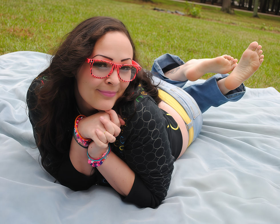 Woman in red glasses and colorful bracelets lies on a blanket in a grassy park, smiling. She's wearing a black top and jeans.
