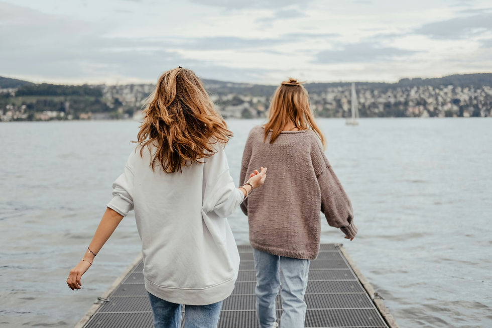 Two people walk on a dock toward a lake, wearing casual sweaters and jeans. A sailboat is visible on the water under a cloudy sky.