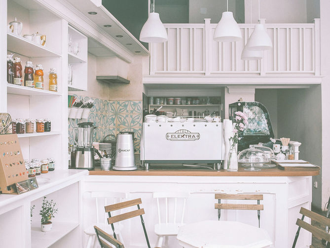 Cozy cafe interior with white decor, light wood chairs, and a counter featuring a white Elektra coffee machine. Warm and inviting ambiance. faith-based family decision. A quiet café breakfast symbolizing a pivotal conversation about faith and family decisions.