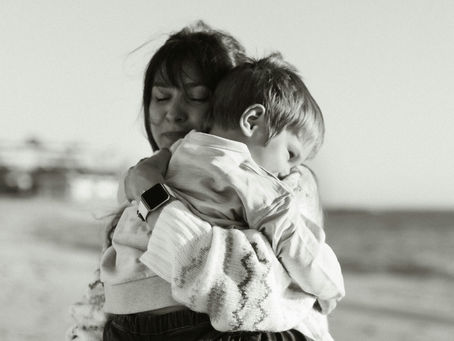 Woman hugs child on beach, both with eyes closed in contentment. She's wearing a sweater and watch. Sand and sea visible in background.