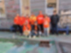 A group of 10 individuals facing the camera for a photo, 9 of whom are wearing orange attire, some holding traditional hand drums in front of a graffiti'd gray wall building