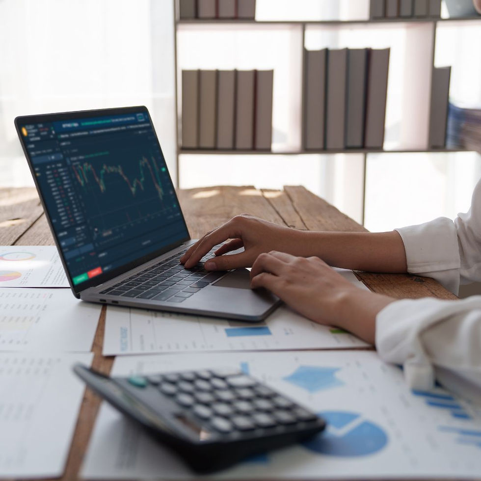 Person working on a laptop displaying financial charts, surrounded by papers and a calculator on a wooden desk. Shelves with books behind.