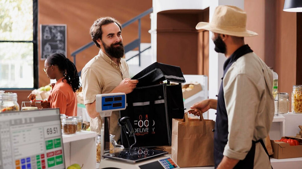 Man in brown shirt receives food delivery bag from shopkeeper in a rustic market. Counter with produce, chalkboard, and digital scale visible.