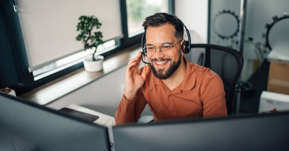 Man with glasses and beard wearing a headset, smiling at a computer in an office with a potted plant by the window.