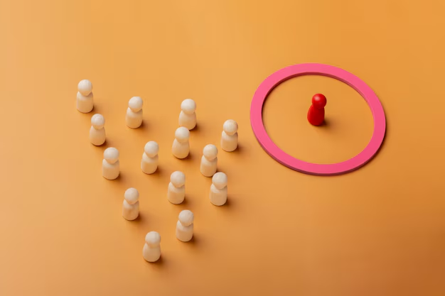 Pawns on orange surface, one red pawn isolated in a pink circle, and a group of white pawns facing it in formation, symbolizing exclusion.