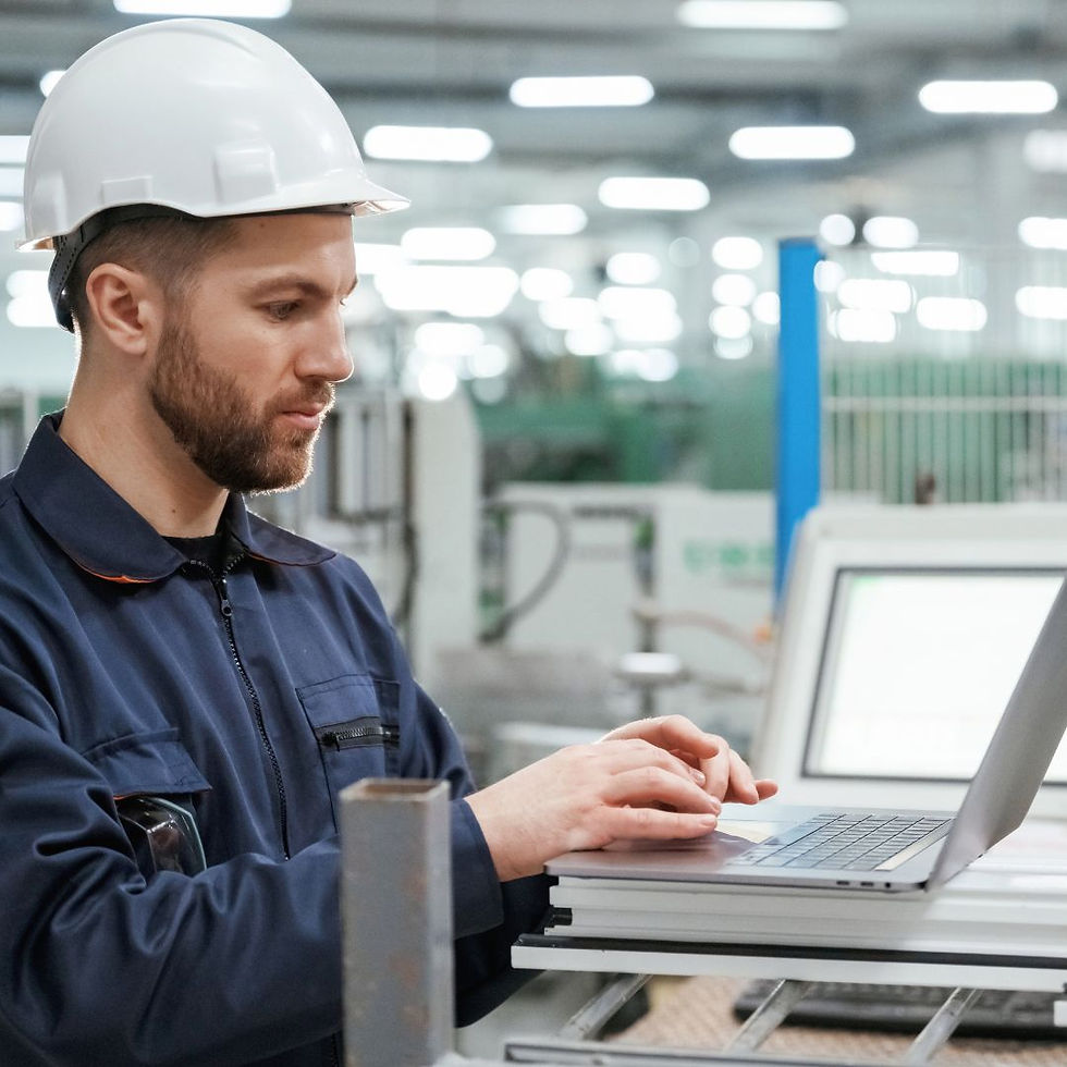 Man in hard hat using laptop in bright factory. Wearing blue coveralls, focused on work. Industrial background with machines.