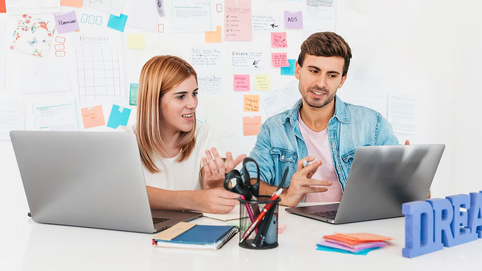 Two people discussing work at a desk with laptops, colorful sticky notes on wall. Pens in holder, books, papers present. Text: "DREAM".