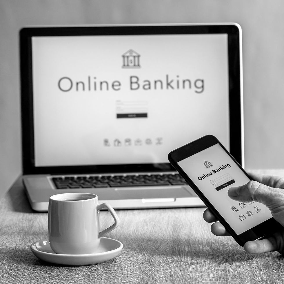 Laptop and phone showing "Online Banking" screen. A hand holds the phone. A coffee cup is on the table. Black and white setting.