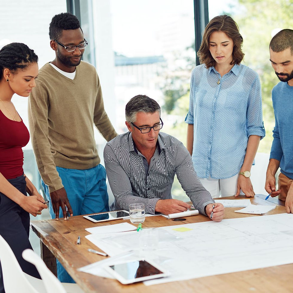 Five people in an office discuss blueprints on a wooden table. Natural light from large windows. Serious and focused mood.