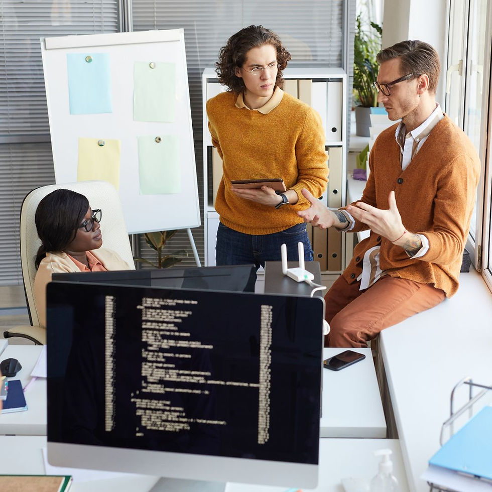 Three people in an office discuss code on a screen. A woman sits, while two men in orange sweaters stand and talk. Neutral colors dominate.