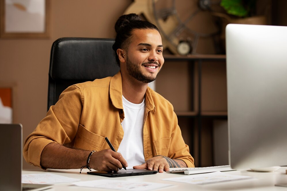 A person smiling while sitting at a desk and working with a graphic tablet, surrounded by design materials and a desktop computer.