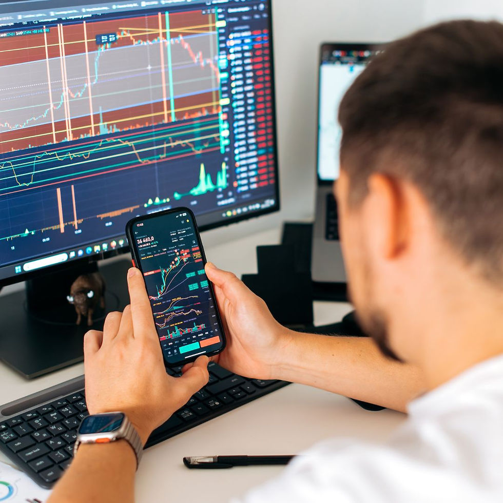 Person checking stock charts on phone and monitor. Desk with keyboard, Apple Watch visible. Trading environment, analytical mood.
