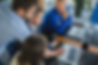 Four people collaborate at a table with laptops, one pointing at a screen. They're in a modern office with a plant visible. Mood is focused.