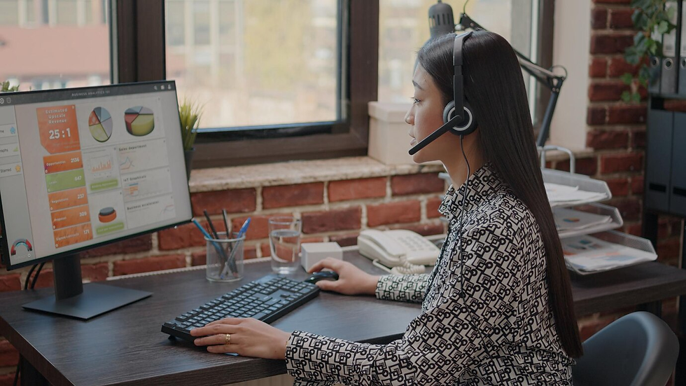 Woman in headset working at a desk with a computer screen displaying colorful charts. Brick wall and window in the background.