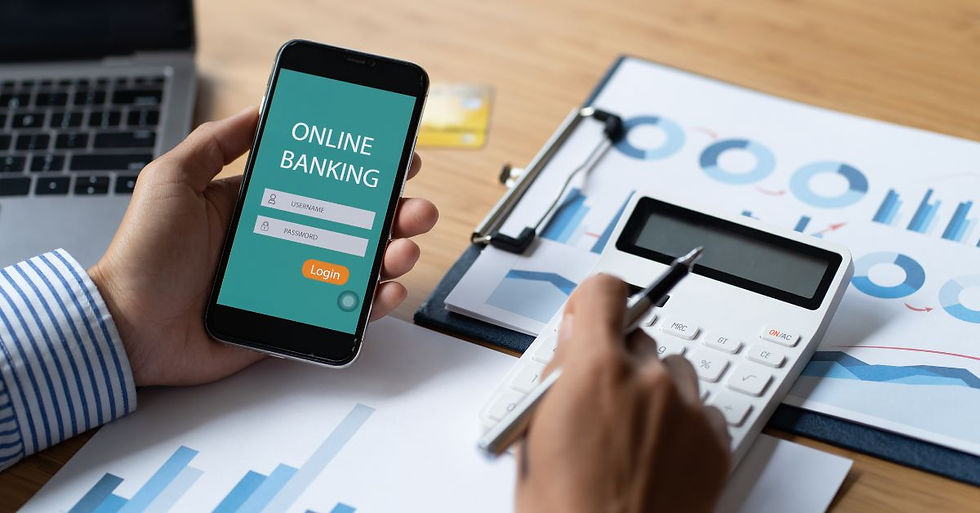 Person holding phone with "Online Banking" login screen, writing on calculator on desk with charts, laptop, and credit card. Business setting.