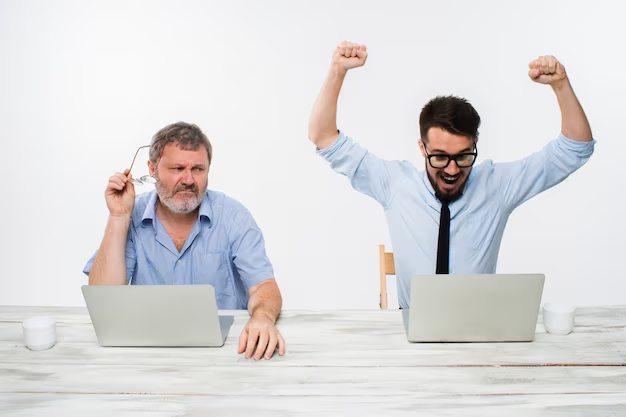 Two men sitting at a table with laptops. One looks frustrated, holding glasses. The other is cheering, arms raised. Both wear blue shirts.