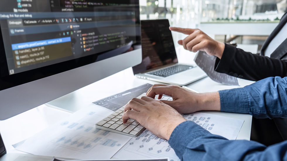 Hands typing on a keyboard with code on a monitor and a person pointing. Papers scattered on a desk. Bright, modern office setting.