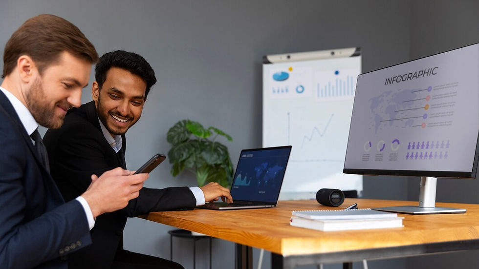Two men in suits smiling at a phone and laptop in an office. A monitor shows an infographic. A plant and chart are in the background.