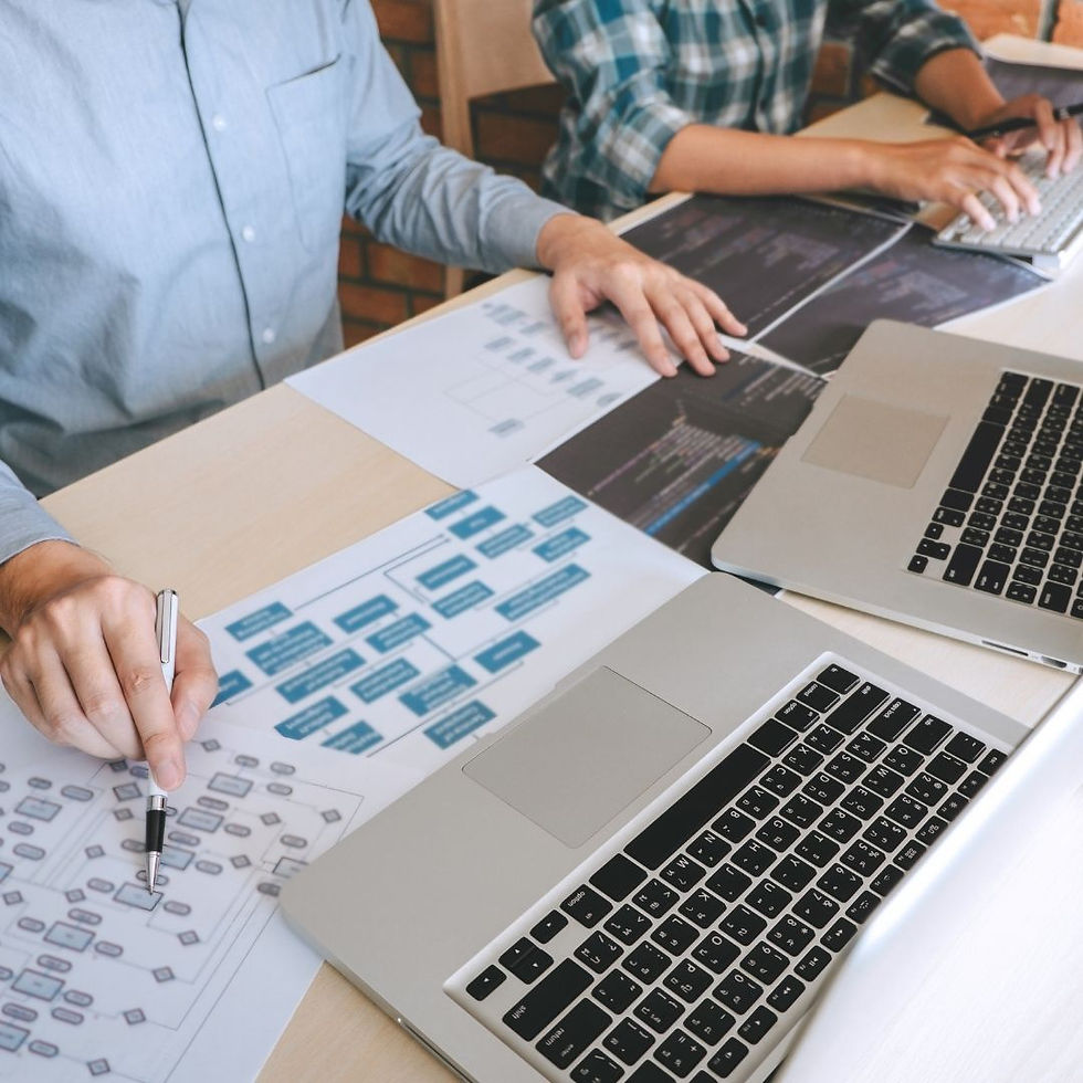 Two people working at a desk with laptops, analyzing flowcharts on papers. Casual setting, brick backdrop, focused atmosphere.
