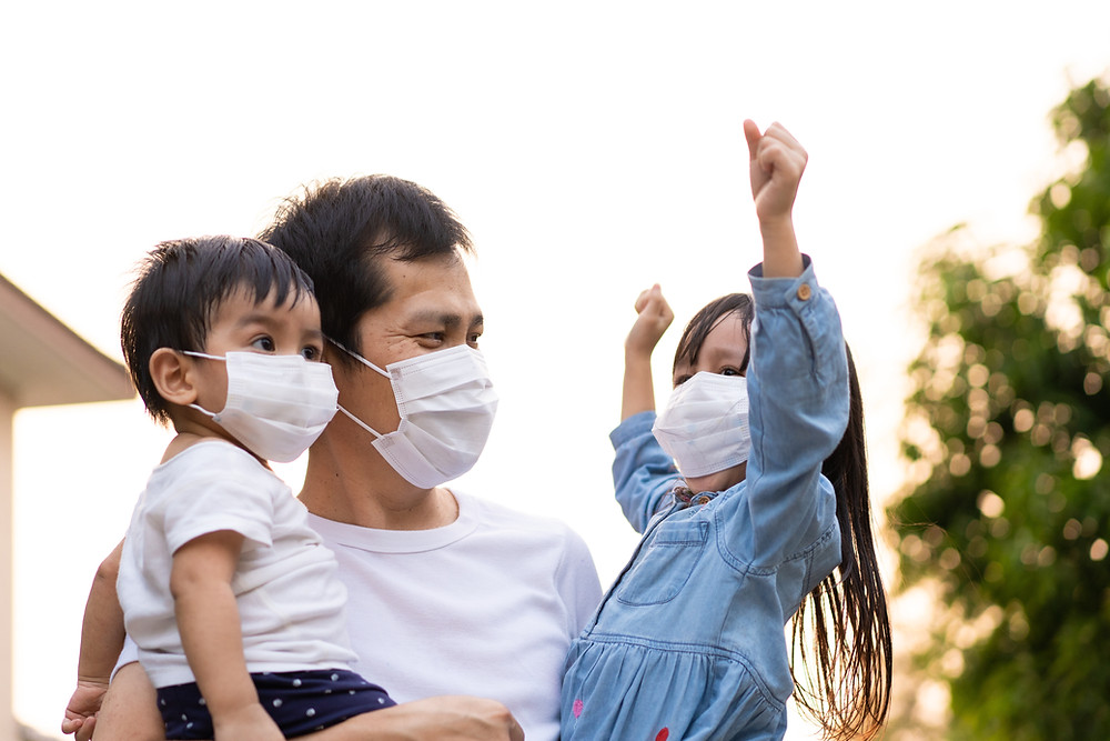 A father holds his two children, all wearing masks, on a sunny day