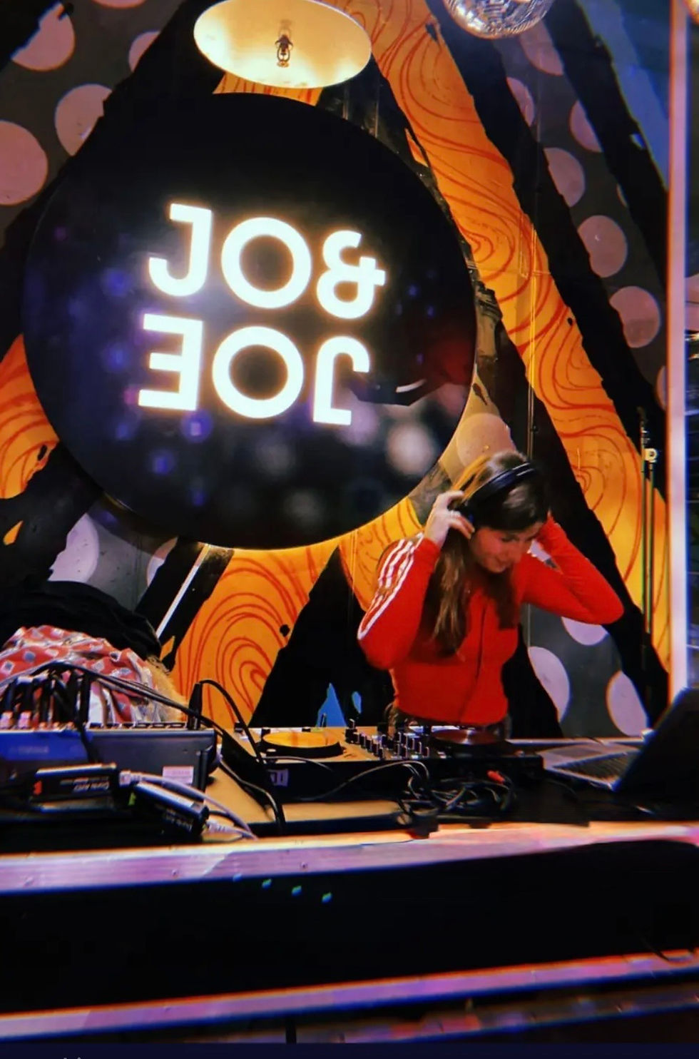 female DJ in a red jacket mixes music with headphones on. Vibrant swirled wall pattern, visible “JOE & JO” text, captures energetic mood.