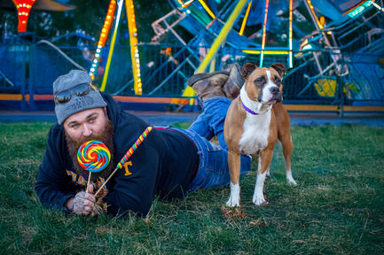 Man at amusement park eating giant, colorful sucker , as he lays on the ground, with dog standing beside him.