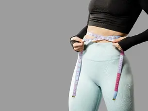A woman with a measuring tape in her hand standing against a grey wall in yoga outfit 
