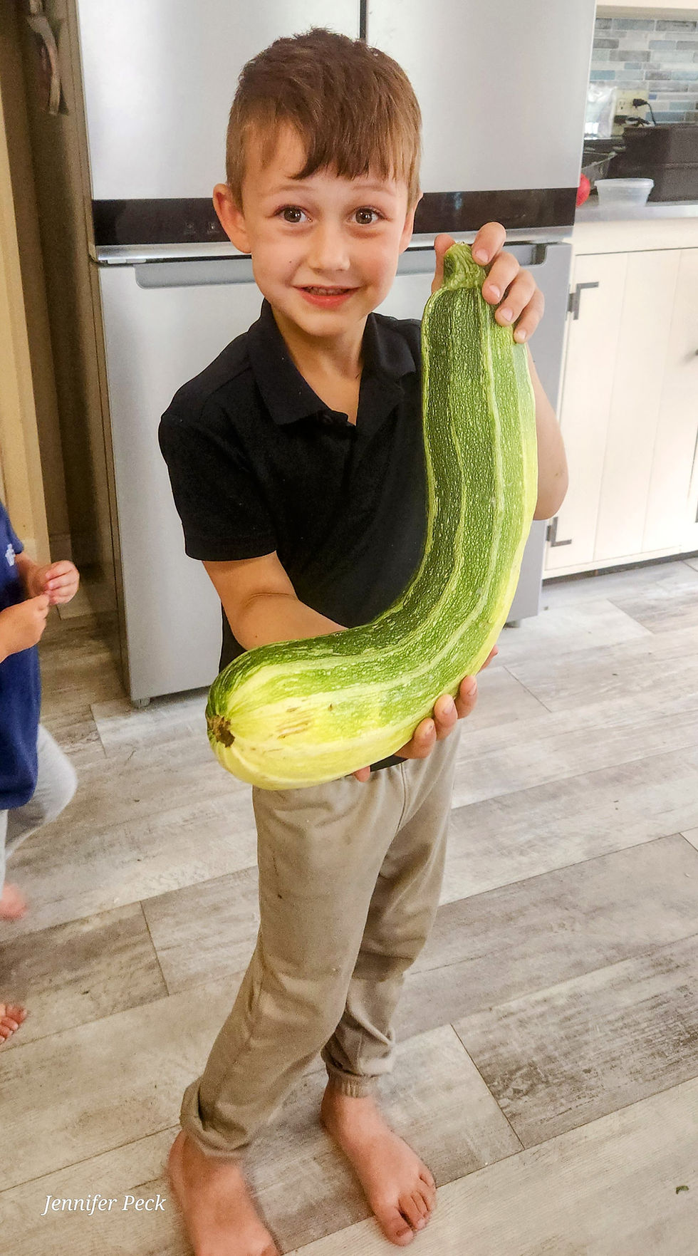 Jackson stands barefoot in a kitchen, smiling at the camera while holding an oversized curved green zucchini with both hands.
