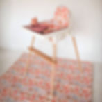 A baby high chair with a tray and wooden legs. The high chair is on a floral patterened splash mat.