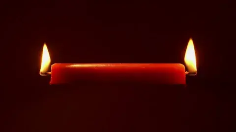A red candle burns at both ends against a dark background, creating a warm glow. The mood is intense and dramatic.