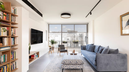 סלון בדירת מגורים בתל אביב עם ספריית עץ פתוחה, ספה אפורה ורצפת בטון בהיר בעיצוב אדריכלי נקי