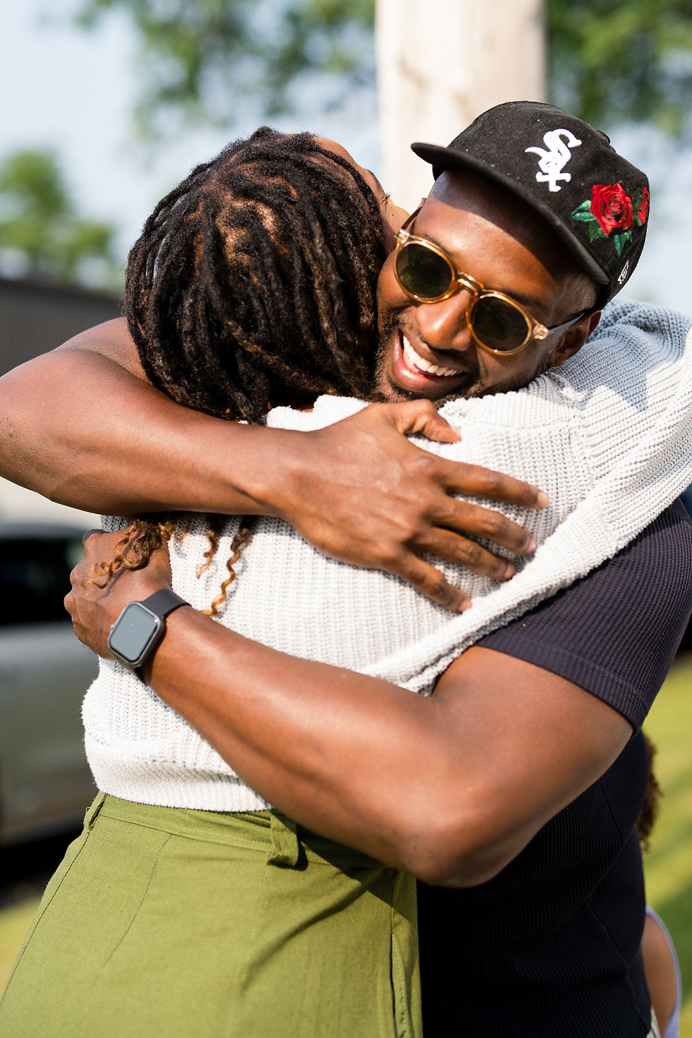 Two people hugging tightly outdoors, one smiling with sunglasses and a baseball cap, showing a joyful expression