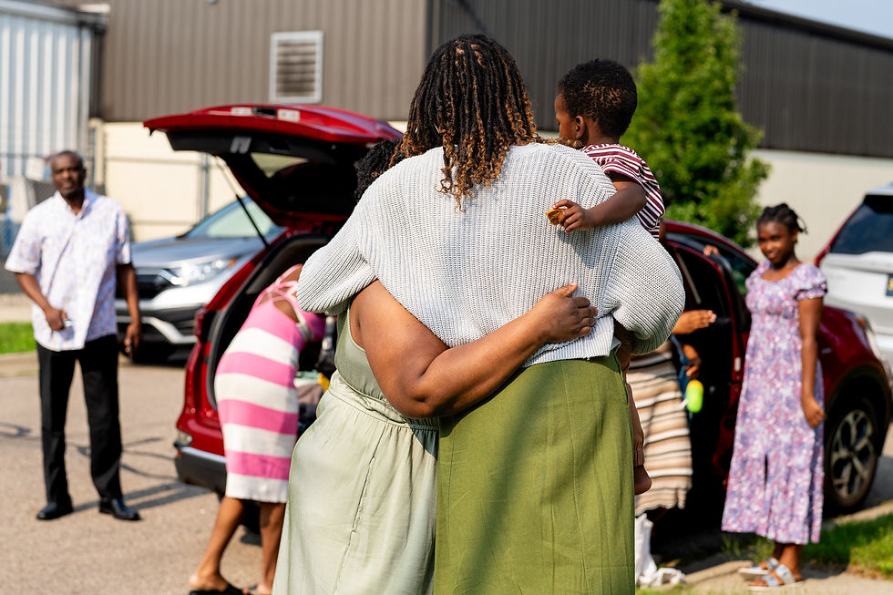Two people hugging while one holds a child, with other family members standing and unloading a car in the background