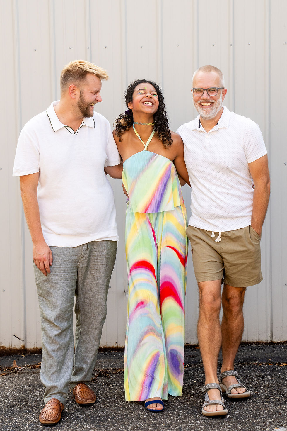 Three people standing together and smiling, with one person in a colorful rainbow dress laughing between two men wearing white shirts
