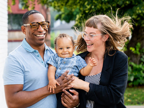 Smiling couple holding a baby outdoors, with the woman’s hair blowing in the wind and trees in the background