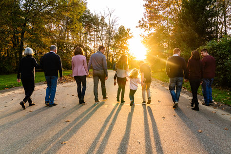 Multi-generational family walking side by side down a paved path toward the sunset, casting long shadows across the road in a tree-lined park