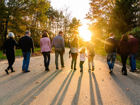 Multi-generational family walking side by side down a paved path toward the sunset, casting long shadows across the road in a tree-lined park