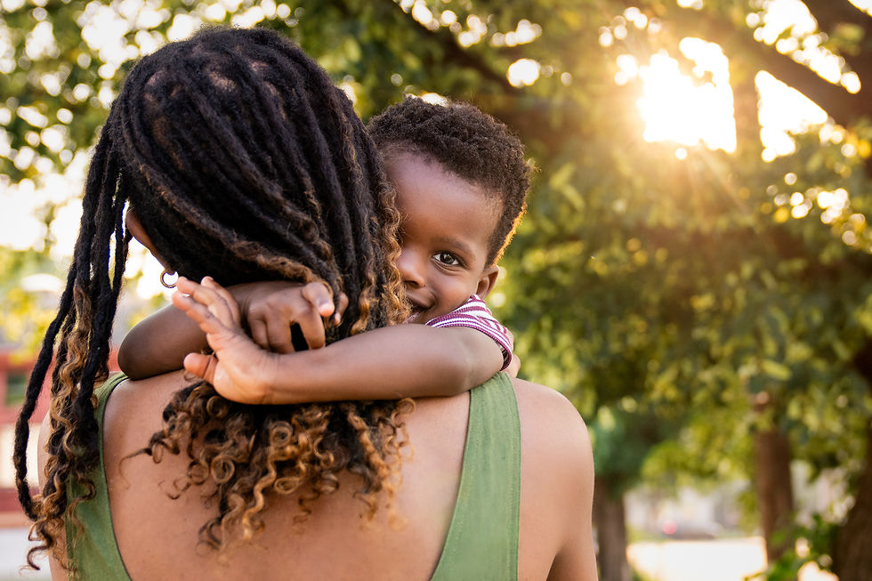 Child resting on a woman’s shoulder, peeking over with a soft smile while embraced in golden evening sunlight under green trees
