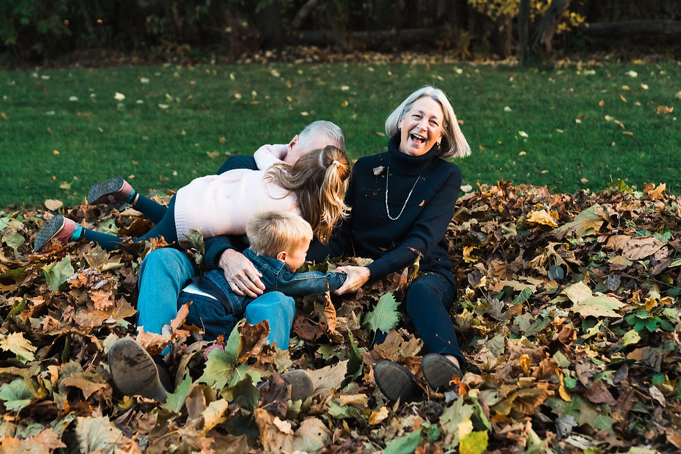 Grandparents and grandchildren laughing and playing in a large pile of autumn leaves, with one child jumping into the pile and everyone smiling in the golden afternoon light