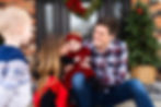 A mother and father smiling as they hold their baby dressed in a red velvet holiday outfit, capturing a festive family moment on the front porch