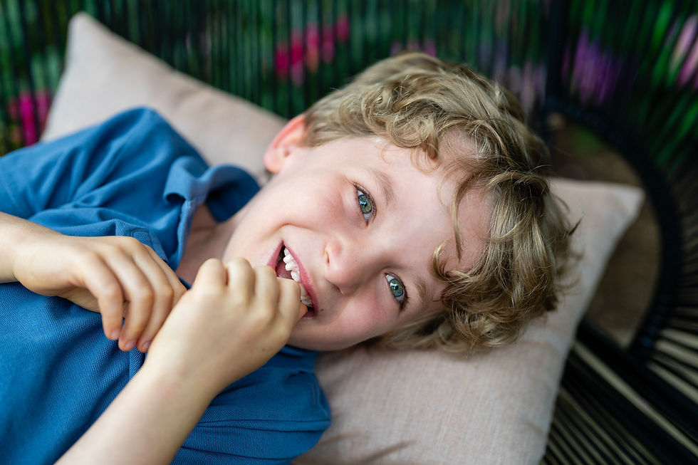 Close-up of a smiling young boy with curly blonde hair and blue eyes, lying on an outdoor pillow and looking up with a playful expression