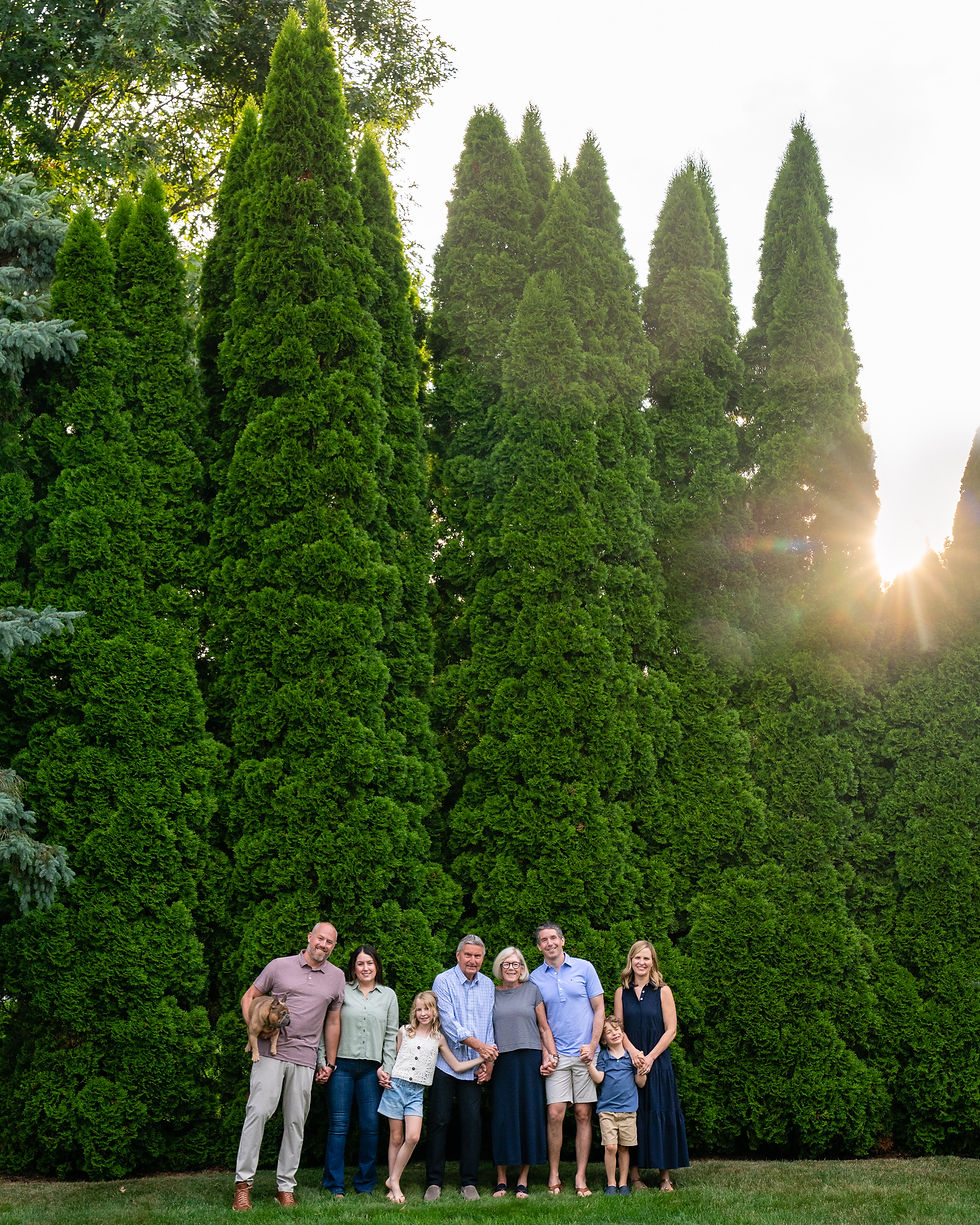 Extended family standing together in front of tall green arborvitae trees at sunset, smiling and holding hands, with a warm sunburst peeking through the trees