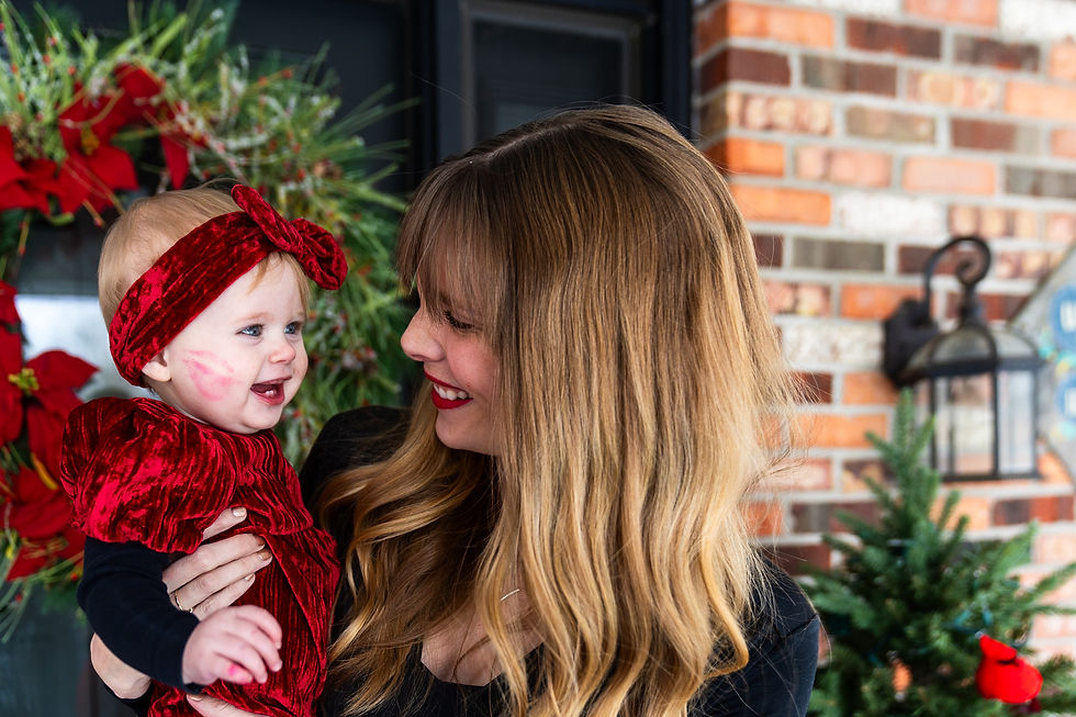Smiling mother holding her baby girl dressed in red velvet with a matching headband, both laughing together in front of a festive Christmas wreath and holiday greenery