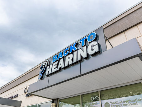 Sign reading "Back to Hearing" on a building under a cloudy sky. Storefront advertises hearing services like hearing test and earwax removal.