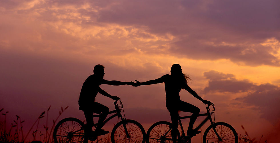 Two people ride bikes holding hands against an orange sunset. Silhouettes are prominent, creating a peaceful, romantic mood.