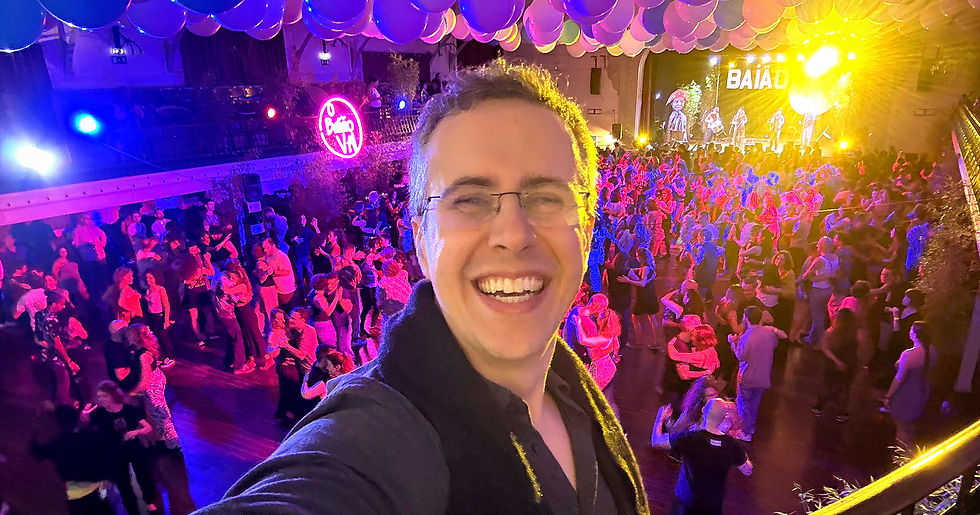 Selfie taken from a dance floor at a forró festival, with the photographer facing away from the crowd. Behind him, a large group of people dance under red, pink, and blue lighting, with a stage visible in the background.