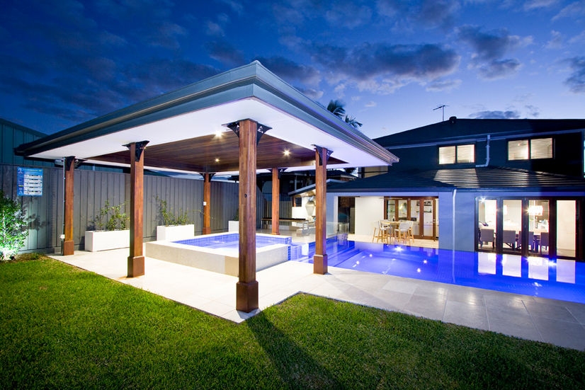 Evening view of luxury backyard featuring ambient lighting, glass pool fence, timber spa surround, and statement planting.