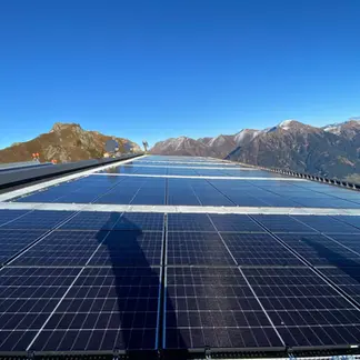 Photovoltaikanlage in den Alpen mit metallische Gitterrost-Wartungswege im Vordergrund. Die Solarpanels erstrecken sich vor einem Panorama aus schneebedeckten Alpen unter strahlend blauem Himmel.